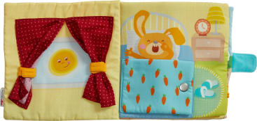 Eine Kinderbuchseite aus Stoff zeigt links eine Sonne mit einem Gesicht hinter roten Vorhängen und rechts einen orangefarbenen Hasen, der im Bett aufwacht, mit Karotten auf der Decke und einem Wecker auf einem Nachttisch.