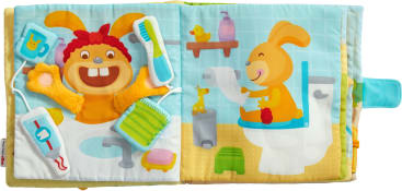 Ein Kinderbuch aus Stoff zeigt illustrierte Kaninchen in einem Badezimmer. Eine Seite zeigt ein lächelndes Kaninchen mit Zahnbürste, Zahnpasta und Seife. Die andere Seite zeigt ein Kaninchen, das auf einer Toilette sitzt und Toilettenpapier hält.