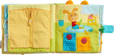 Ein Kinderbuch aus Stoff ist auf zwei Seiten geöffnet. Die linke Seite zeigt eine gelbe Reißverschlusstasche und die rechte Seite zeigt ein Kaninchen, das auf dem Boden spielt, mit zwei Taschen, die mit roten Knöpfen geschlossen werden.