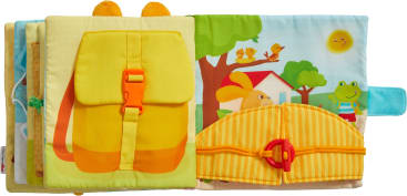 Ein aufgeschlagenes Kinderbuch aus Stoff, das auf der linken Seite einen gelben Rucksack mit einer orangefarbenen Schnalle und auf der rechten Seite eine gelb gestreifte, halbkreisförmige Klappe mit einem roten Clip zeigt; im Hintergrund ist eine Zeichentrickszene im Freien zu sehen.