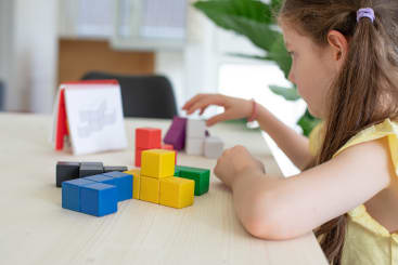 Ein junges Mädchen sitzt an einem Tisch und ordnet bunte geometrische Blöcke an. Eine Anweisungskarte mit einem 3D-Blockmuster liegt auf dem Tisch neben ihr. Im Hintergrund sind grüne Blätter zu sehen.