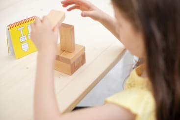 Ein Kind in einem gelben Hemd baut an einem hellen Tisch ein Bauwerk aus Holzklötzen. Ein kleines gelbes Heft mit einem Diagramm liegt aufgeschlagen neben ihnen. Das Gesicht des Kindes ist nicht zu sehen.
