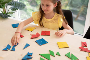 Ein Mädchen in einem gelben Hemd sitzt an einem hellen Holztisch und ordnet bunte geometrische Puzzleteile in verschiedenen Formen an. Im Hintergrund ist ein großes Fenster mit Grünpflanzen zu sehen.