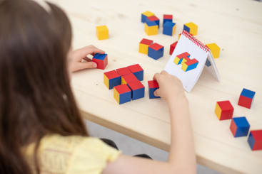 Ein Kind sitzt an einem Tisch und ordnet rote, blaue und gelbe Würfel so an, dass sie einem 3D-Blockmuster entsprechen, das auf einem kleinen Daumenkino vor ihm abgebildet ist. Mehrere farbige Würfel sind auf der hellen Holzoberfläche verstreut.