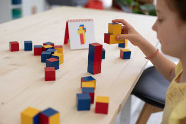 Ein Kind sitzt an einem Tisch und stapelt rote, gelbe und blaue Holzwürfel. Eine Staffelei mit einer Karte, die eine Figur aus ähnlichen Würfeln zeigt, steht auf dem Tisch zwischen verstreuten Blöcken.