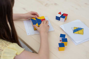 Ein Kind ordnet farbige Würfel mit roten, blauen und gelben Flächen so an, dass sie einem geometrischen Muster entsprechen, das auf einer Karte auf einem hellen Holztisch abgedruckt ist. Andere Musterkarten und Würfel liegen in der Nähe.