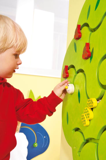 Ein kleines Kind in einem roten Pullover bewegt ein kleines Objekt auf einer grünen, baumförmigen Aktivitätswand mit roten und gelben Teilen. Im Hintergrund sind weitere bunte Wanddekorationen zu sehen.