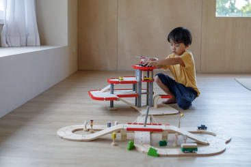 Ein kleines Kind in einem gelben Hemd und einer blauen Hose sitzt auf einem Holzboden und spielt in einem hellen, minimalistischen Raum mit einer Spielzeug-Parkgarage und einem hölzernen Autorennbahn-Set.