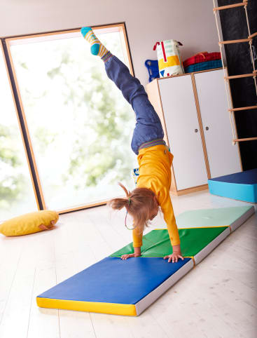 Ein Kind in einem gelben Hemd und einer blauen Hose macht einen Handstand auf einer gepolsterten Matte in einem hellen Raum mit Holzboden, einem großen Fenster, Schränken und einem gelben Kissen auf dem Boden.
