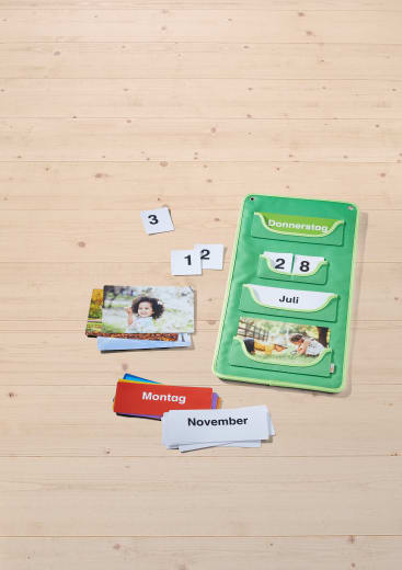 Ein grüner Organizer mit Kartenfächern zeigt "Donnerstag", "28" und "Juli" an. In der Nähe befinden sich Zahlenkarten, Karten mit Tages- und Monatsnamen, Fotokarten und eine Karte mit der Aufschrift "Montag". Alle befinden sich auf einer hellen Holzoberfläche.