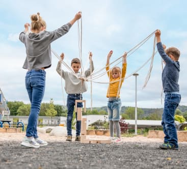 Vier Kinder stehen im Freien in einem Kreis und halten jeweils Schnüre, die an einer Holzstruktur auf dem Boden befestigt sind. Sie heben ihre Arme und ziehen die Schnüre gemeinsam hoch. Im Hintergrund sind Spielplatzgeräte und Gebäude zu sehen.