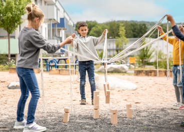 Drei Kinder stehen im Freien auf einem Spielplatz und halten jeweils Seile, die mit Holzklötzen verbunden sind. Die Kinder arbeiten zusammen, um die Blöcke zu stapeln, während im Hintergrund Gebäude und Grünflächen zu sehen sind.
