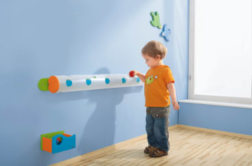 Ein kleines Kind in einem orangefarbenen Hemd steht in einem Raum und legt einen Ball in eine transparente horizontale Röhre mit blauen Punkten an einer hellblauen Wand. Der Raum hat einen hellen Holzboden und Wanddekorationen in Form von Vögeln.
