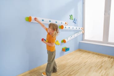 Ein kleines Kind legt einen Ball in eine von drei horizontalen Plastikröhren, die an einer hellblauen Wand angebracht sind. Das Kind hält mehrere Bälle und steht auf einem hellen Holzboden in der Nähe eines Fensters.