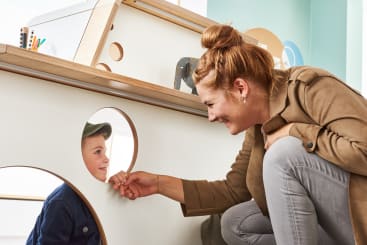 Eine Frau hockt neben einem Spielgerät und lächelt ein kleines Kind an, das sie durch einen runden Ausschnitt in der Platte ansieht. Das Kind trägt eine blaue Jacke und eine grüne Mütze; die Umgebung scheint ein Innenraum zu sein.