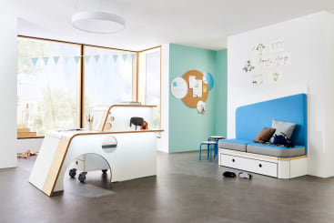 Ein modernes Klassenzimmer mit einem gebogenen Schreibtisch auf Rädern, einer blauen gepolsterten Bank mit Schubladen, einem kleinen blauen Hocker, einer runden Korkplatte, hellen Wänden, bodentiefen Fenstern und Dekoration wie Wandkunst und Wimpeln.