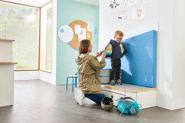 Eine Frau kniet sich hin und hilft einem kleinen Jungen, seine bunte Jacke in einem hellen, modernen Raum zu schließen. Der Junge steht auf einer gepolsterten Bank und lächelt. In der Nähe liegen ein grüner Helm und ein blauer Rucksack auf dem Boden.