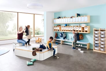 Ein Klassenzimmer mit vier kleinen Kindern und einem Erwachsenen. Die Kinder spielen und interagieren miteinander. Der Raum hat Regale mit Fächern, Behältern, Jacken und Rucksäcken. Große Fenster lassen natürliches Licht herein.