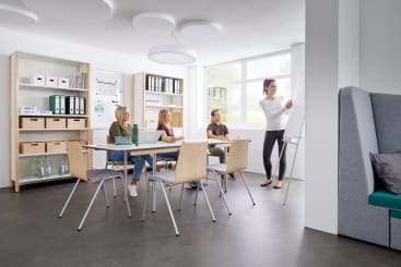 Eine Frau steht an einem Flipchart und hält eine Präsentation vor drei sitzenden Kollegen in einem modernen Besprechungsraum mit Regalen, Dokumenten und Laptops.