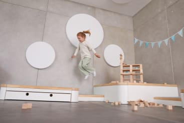 Ein kleines Kind mit roten Haaren hüpft in einem modernen Spielzimmer mit grauen Wänden, weißen runden Wanddekorationen, Holzklötzen und einer kleinen Holzkonstruktion. Einige Klötze liegen auf dem Boden verstreut. Blaue Wimpel hängen an der Wand.