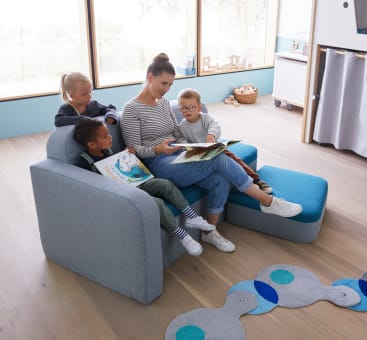 Ein Erwachsener sitzt mit drei kleinen Kindern auf einer modernen blauen Couch und liest ein Buch. Die Kinder sitzen dicht beieinander und schauen sich die Seiten an. Der Raum ist hell mit großen Fenstern, Holzboden und Kinderspielzeug in der Nähe.