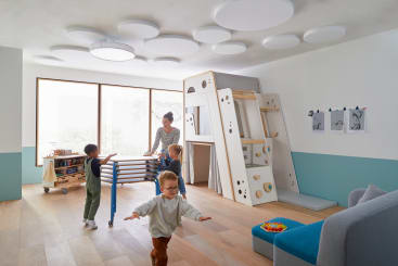 Drei kleine Kinder spielen in einem hellen, modernen Spielzimmer mit einem Klettergerüst und einer Rutsche. Ein Erwachsener steht in der Nähe neben einem Rollwagen. Kunstzubehör und Zeichnungen sind zu sehen; der Raum hat große Fenster und Holzböden.