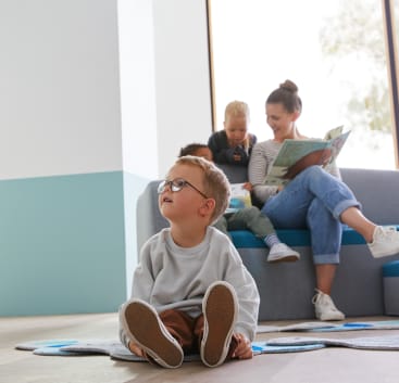 Im Vordergrund sitzt ein kleines Kind mit Brille auf dem Boden, während im Hintergrund eine Frau zwei Kindern auf einer Couch vorliest. Der Raum hat hellblaue und weiße Wände und große Fenster.