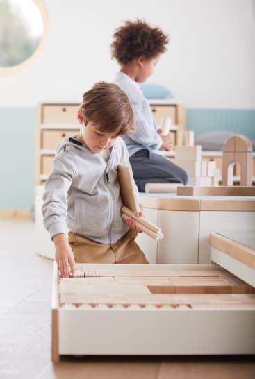 Zwei kleine Kinder spielen mit Holzklötzen in einem hellen, modernen Klassenzimmer. Ein Kind wählt Blöcke aus einer Schublade aus, während das andere im Hintergrund sitzt und Bausteine hält.