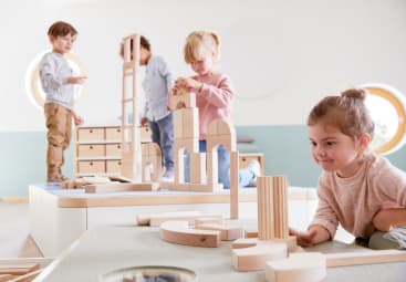Vier kleine Kinder spielen zusammen in einem hellen Raum und bauen mit Holzklötzen hohe Strukturen. Sie sind konzentriert bei der Sache, und auf den Tischen um sie herum sind verschiedene Klötzchentürme zu sehen.