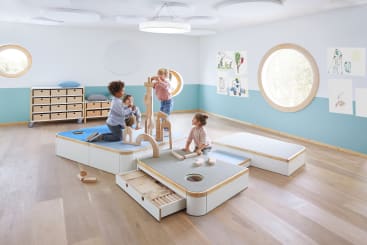 Vier kleine Kinder spielen mit Holzbausteinen in einem hellen, modernen Klassenzimmer mit runden Fenstern, hellen Holzböden und Kinderzeichnungen an den Wänden. Die Kinder sind um modulare Spielmöbel versammelt.