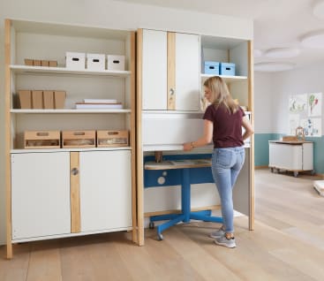 Eine Frau steht in einem Raum neben einer Reihe von Regalen und Schränken und zieht eine weiße Rolltür herunter, um einen Arbeitsbereich auf einem blauen Tisch freizugeben. In den Regalen stehen Aufbewahrungsboxen und Tabletts. Der Boden ist aus hellem Holz.