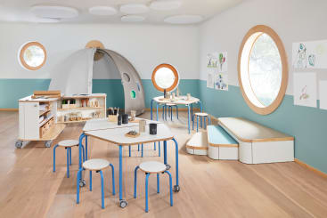 Ein modernes Klassenzimmer mit Holzböden, runden Fenstern, kindgerechten Tischen und Hockern, einer Leseecke, einer Kunststation, einem kuppelförmigen Spielgerät und Kinderkunstwerken an den Wänden.