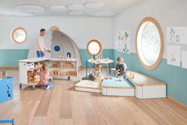 Ein Klassenzimmer mit vier kleinen Kindern, die mit Holzklötzen spielen, und einem Mann, der sie beaufsichtigt. Der Raum hat runde Fenster, Wandmalereien und helle Holzböden. In einer Ecke steht eine graue zeltartige Struktur.