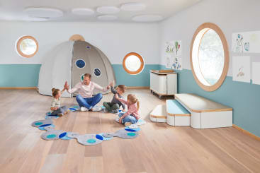 Ein Erwachsener und drei kleine Kinder sitzen im Kreis auf einem gemusterten Teppich in einem hellen Raum mit Holzfußboden, runden Fenstern, einem kuppelförmigen Spielzelt und Kinderkunstwerken an den Wänden.