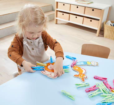 Ein kleines Kind sitzt an einem hellblauen Tisch und befestigt bunte Plastikwäscheklammern an Tierausschnitten aus Papier. Weitere Wäscheklammern liegen verstreut auf dem Tisch. Im Hintergrund sind eine Reihe von Schubladen und Stufen zu sehen.