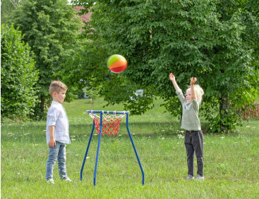 Zwei kleine Kinder spielen im Freien Basketball; ein Kind wirft einen grünen und gelben Ball in einen kleinen Korb, während das andere zusieht. Sie stehen auf einer Wiese mit Bäumen im Hintergrund.
