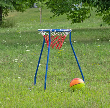 Ein kleiner Basketballkorb mit rotem Netz steht im Gras, daneben liegt ein gelber und oranger Basketball. Im Hintergrund sind Bäume und Wildblumen zu sehen.