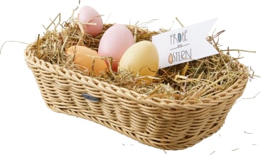 In einem mit Stroh gefüllten Weidenkorb befinden sich vier pastellfarbene Eier und eine weiße Karte mit der Aufschrift "Frohe Ostern".