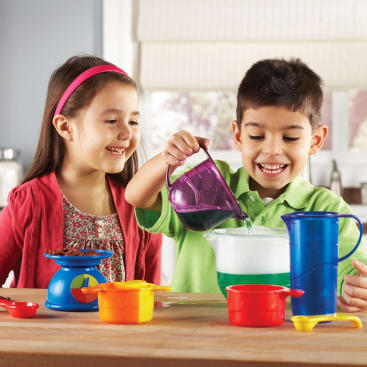 Zwei Kinder spielen mit bunten Plastikmessbechern und einer Waage und gießen Wasser in einen durchsichtigen Behälter auf einer Küchenzeile. Beide lächeln und konzentrieren sich auf ihre Tätigkeit.