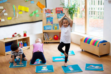 Drei kleine Kinder üben auf einzelnen Matten in einem hellen Klassenzimmer Yogastellungen. Ein Kind steht aufrecht mit den Händen über dem Kopf, während die anderen auf allen Vieren stehen. Im Hintergrund befinden sich Regale und Bücher.