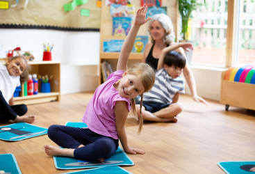 Kinder und ein Erwachsener sitzen auf Matten in einem Klassenzimmer und strecken sich mit einem erhobenen Arm. Das Klassenzimmer hat Holzböden, Bücherregale und bunte Dekorationen. Das Sonnenlicht fällt durch große Fenster ein.