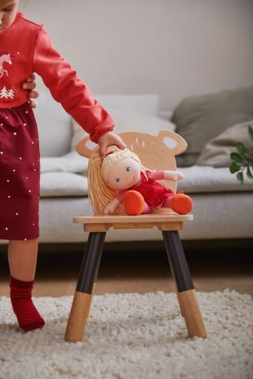 Ein Kind in einem roten Kleid und roten Socken setzt eine blonde Puppe in einem roten Outfit auf einen kleinen Holzstuhl mit tierförmigen Details. Die Szene spielt in einem Wohnzimmer mit einem weißen Teppich und einem Sofa im Hintergrund.