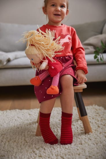 Ein kleines Kind in einem rosa-roten Kleid und roten Socken sitzt auf einem kleinen Holzschemel und hält eine Puppe mit blondem Garnhaar und rotem Outfit. Im Hintergrund sind eine beigefarbene Couch und ein Teppich zu sehen.