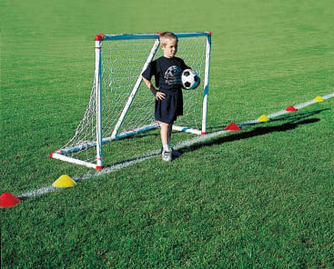Ein kleiner Junge steht vor einem kleinen Fußballtor auf einer Wiese und hält einen Fußball unter einem Arm. Bunte Kegel markieren Linien auf dem Rasen um ihn herum.