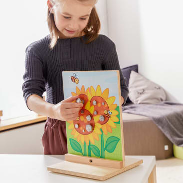 Ein junges Mädchen steht an einem Tisch und setzt runde Holzteile mit Löchern auf ein Brettspiel mit Bienen und Sonnenblumen in einem hellen Raum.
