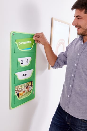 Ein Mann steht neben einem an der Wand befestigten grünen Organizer, der den Tag Donnerstag, das Datum 24 und den Monat Juni anzeigt, zusammen mit einem Foto von zwei Personen an einem Tisch in der unteren Tasche.