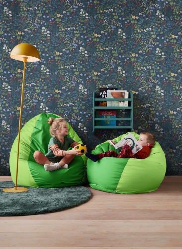 Zwei Kinder sitzen auf hellgrünen Sitzsäcken in einem Raum mit Blümchentapete. Ein Kind hält einen Fußball, während das andere ein Buch liest. Im Hintergrund stehen eine gelbe Stehlampe und ein kleines Regal mit Büchern.