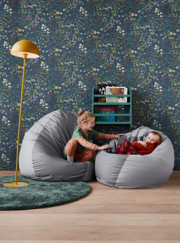 Zwei Kinder sitzen auf großen grauen Sitzsäcken in einem Raum mit einem grün gemusterten Teppich, einer gelben Stehlampe und einem Bücherregal vor einer dunklen Blümchentapete.