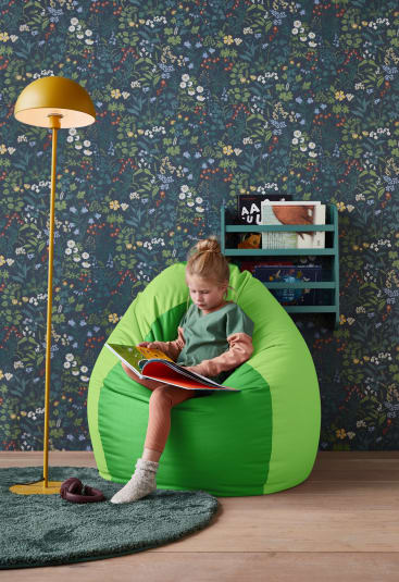 Ein kleines Kind sitzt auf einem grünen Sitzsack und liest ein Buch. Das Zimmer hat eine Blumentapete, eine gelbe Stehlampe, einen grünen Teppich und ein Wandregal mit Büchern und Spielzeug.