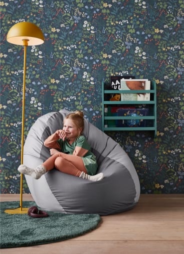 Ein kleines Kind sitzt auf einem grauen Sitzsack in einem Raum mit Blümchentapete, einem grünen runden Teppich, einer gelben Stehlampe und einem an der Wand befestigten Zeitschriftenständer mit Büchern.